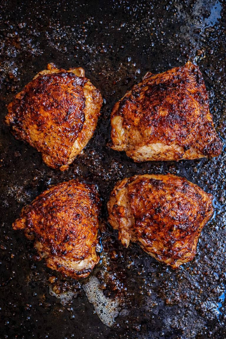 Four deeply browned, caramelized chicken thighs cooking on a dark sheet pan with bubbling glaze and rendered fat.